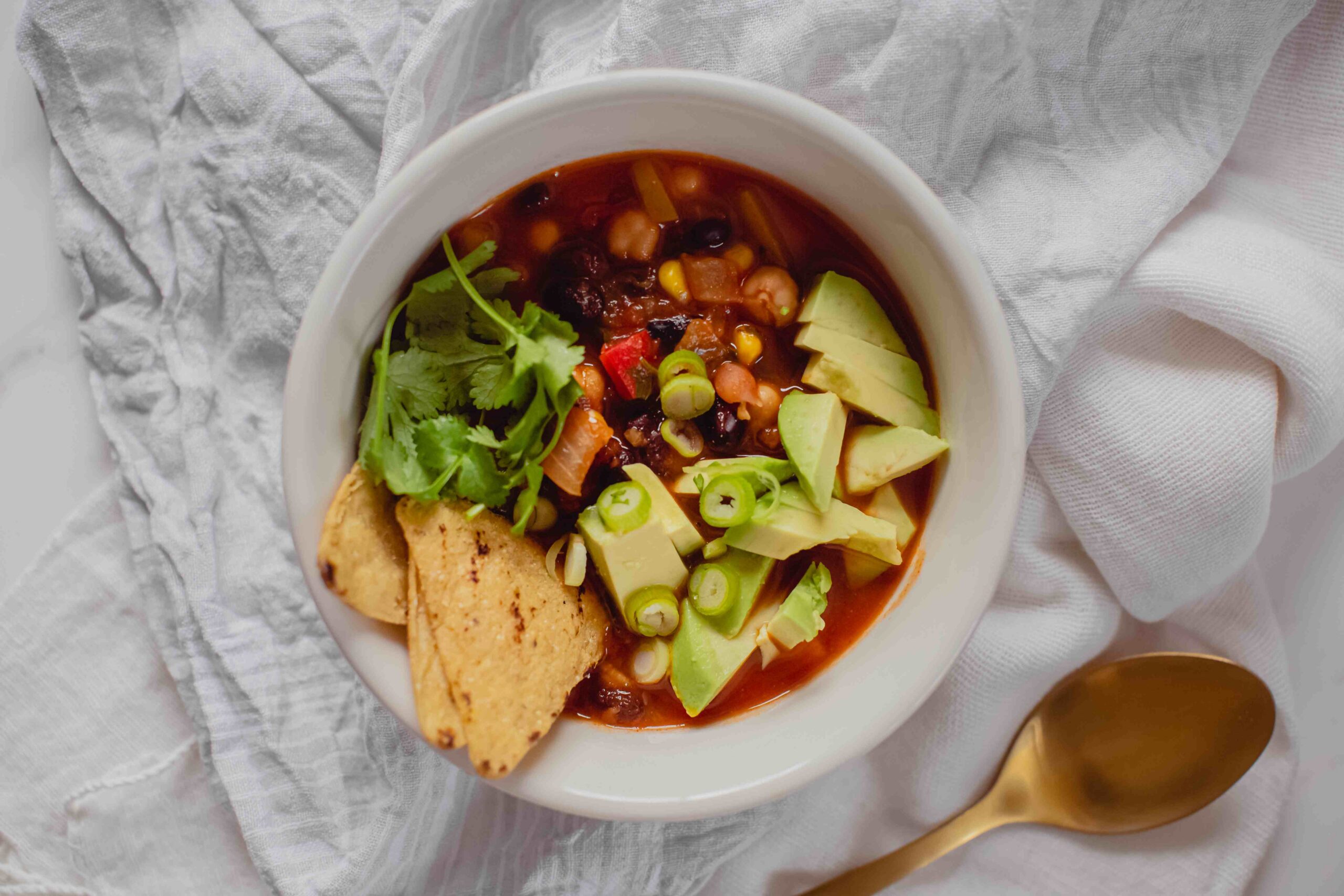 Hero image of a bowl of Mexican Bean Soup topped with corn chips, spring onion, cilantro and avocado, on a white kitchen counter.